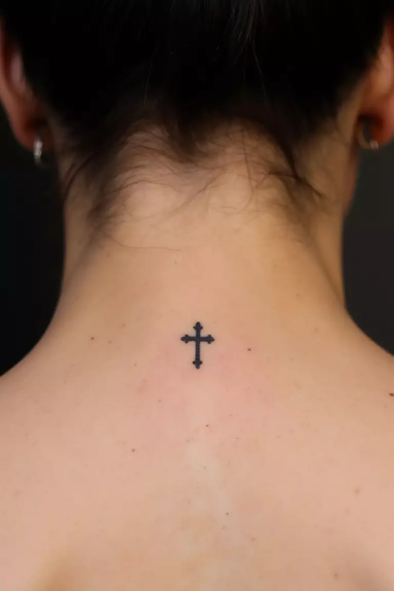 Tiny Cross Tattoo A close up shot of woman's neck from the back with a black ink tiny cross tattoo