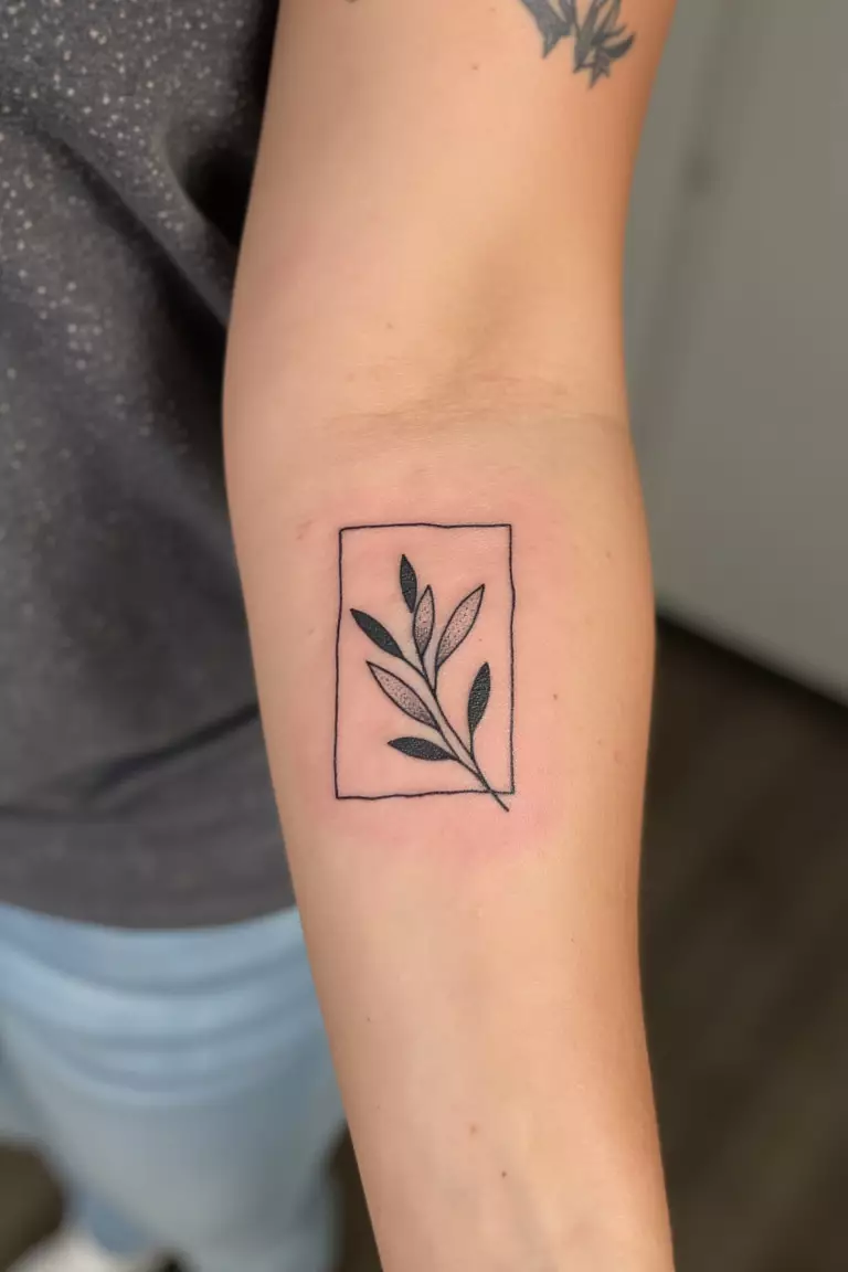 Olive Branch With A Minimalist Frame A close up shot of woman's arm with a black ink olive branch within a minimalist frame tattoo