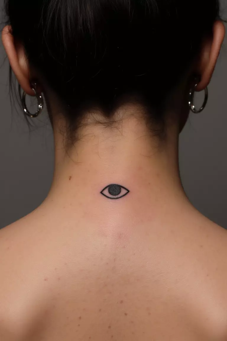 Tiny Evil Eye Tattoo A close up shot of woman's neck from the back with a black ink tiny evil eye tattoo