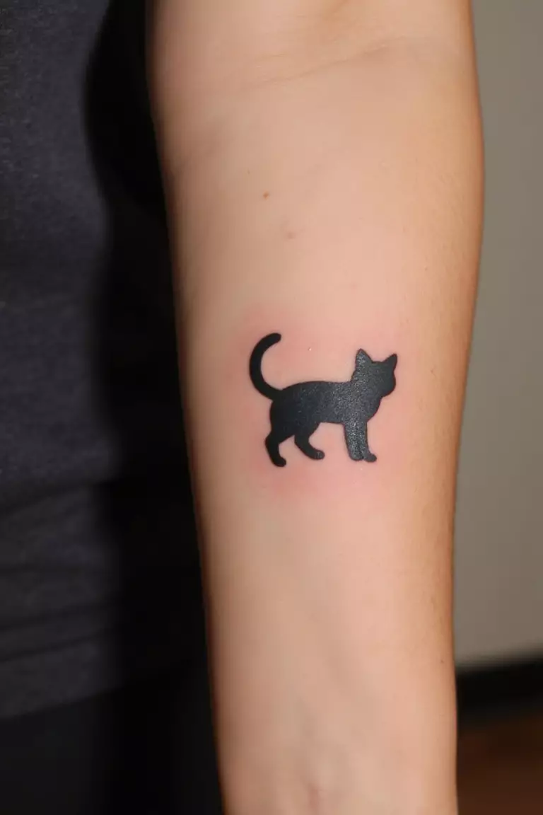 Minimalist Black Cat Tattoo A close up shot of woman's arm with a black ink tattoo of a simple black cat silhouette.