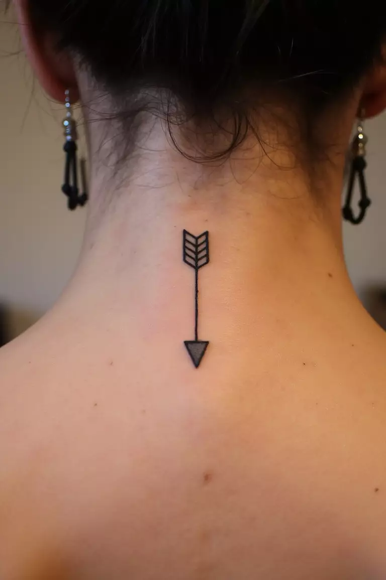 Tiny Arrow Tattoo A close up shot of woman's neck from the back with a black ink tiny arrow tattoo
