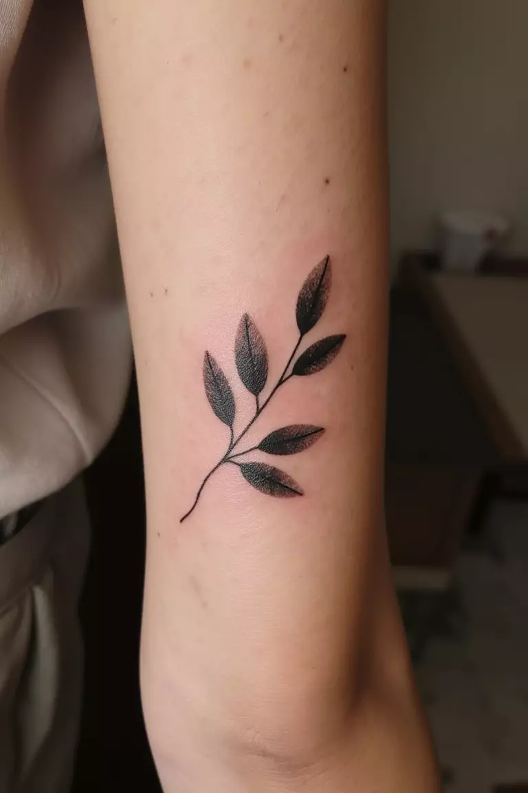 Olive Branch With A Subtle Watercolor Effect A close up shot of woman's arm with a black ink olive branch featuring a subtle watercolor effect tattoo