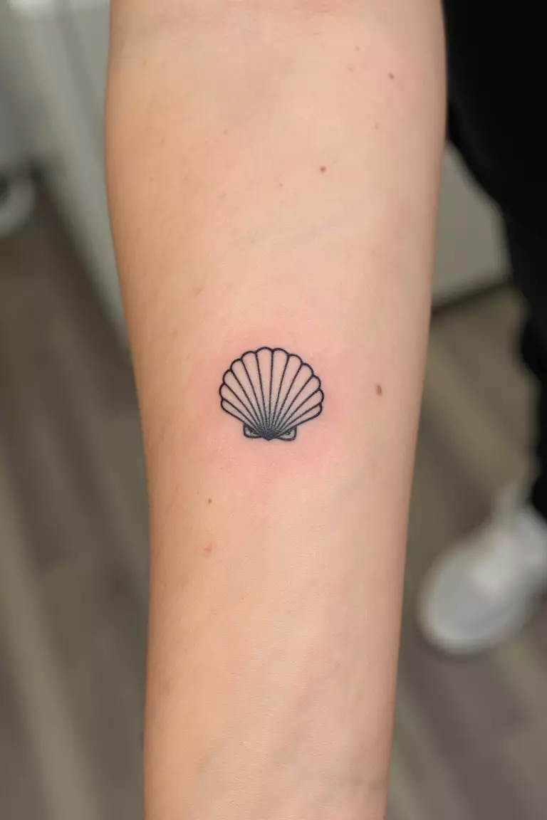 Sea Shell A close up shot of woman's arm with a black ink tattoo showing a tiny seashell with intricate fine line details giving definition, minimalistic beach design.