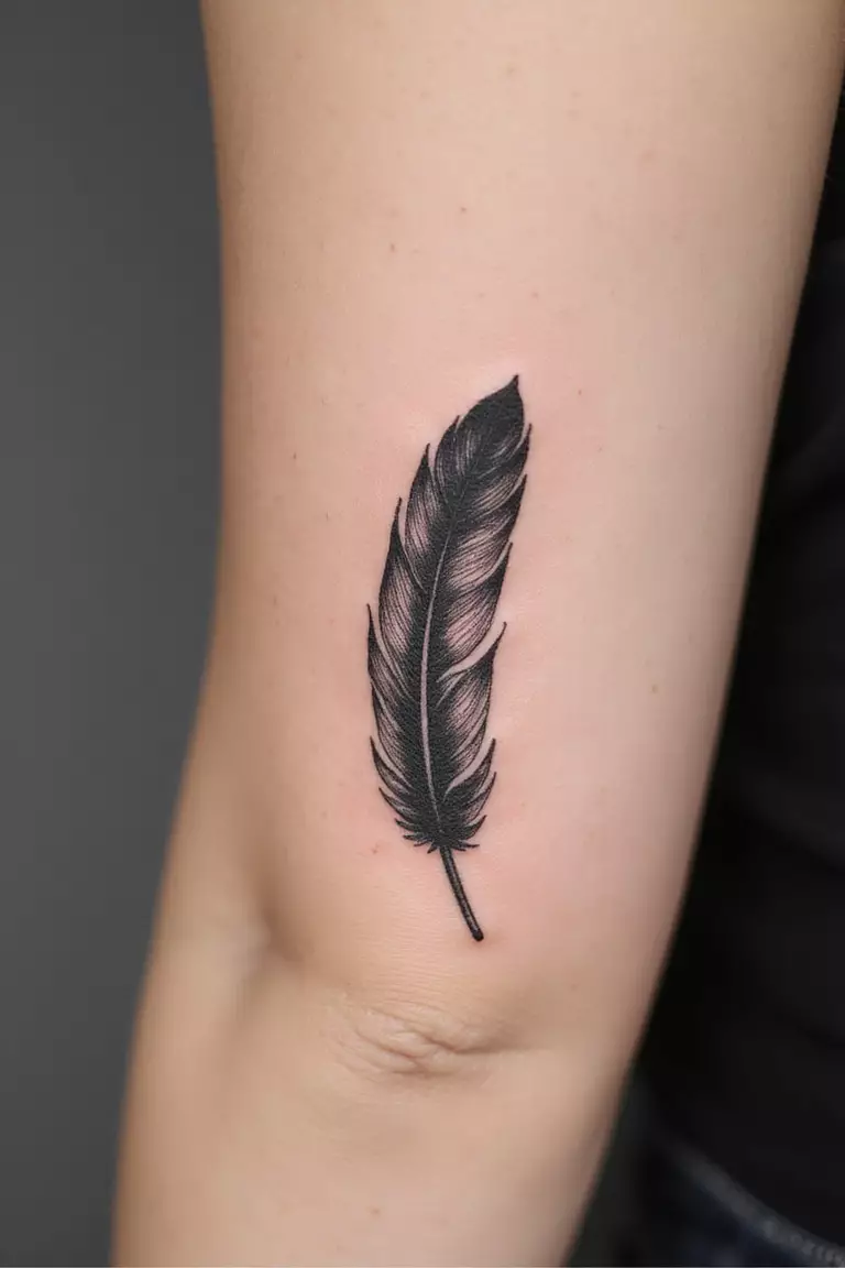 Feather A close-up shot of a woman's arm with a black ink tattoo of a single, detailed feather seemingly floating down.
