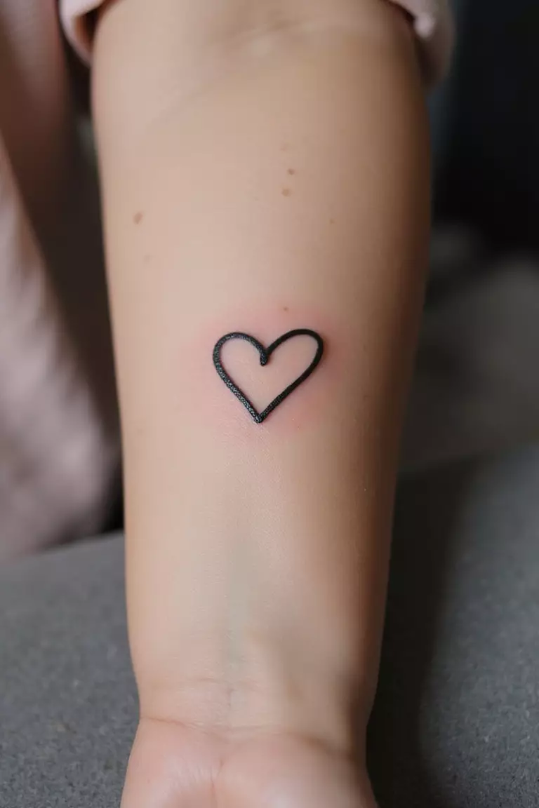 Tiny Heart Outline A close-up shot of a woman's arm with a black ink tattoo of a tiny, simple heart outline.