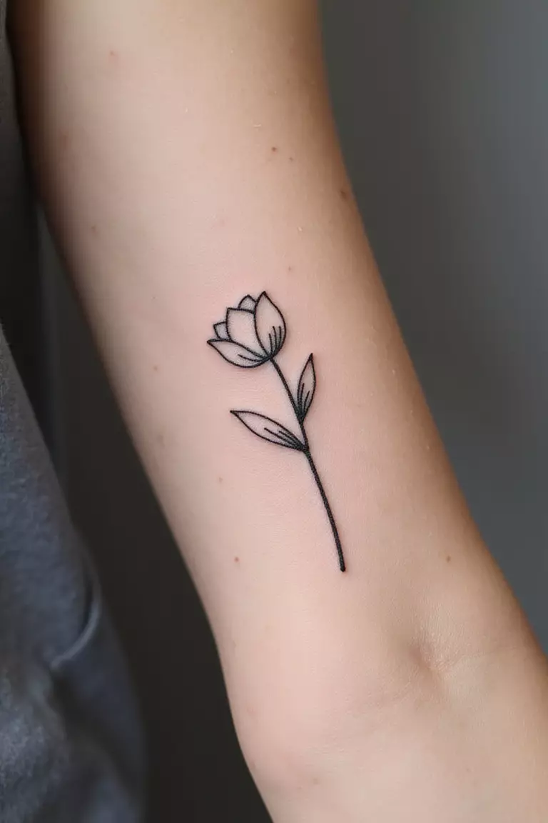 Single Minimalist Flower A close-up shot of a woman's arm with a black ink tattoo of a single stem minimalist line art flower, like a tulip or daisy.
