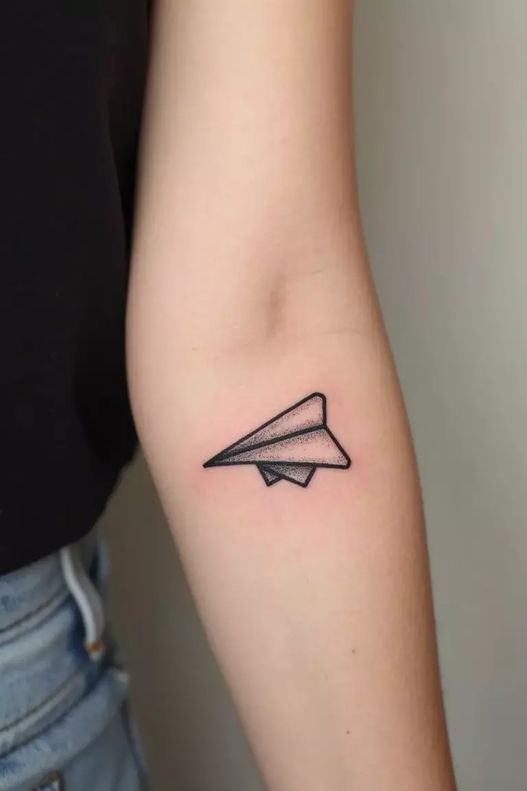Paper Airplane A close-up shot of a woman's arm with a black ink tattoo of a simple line art paper airplane.