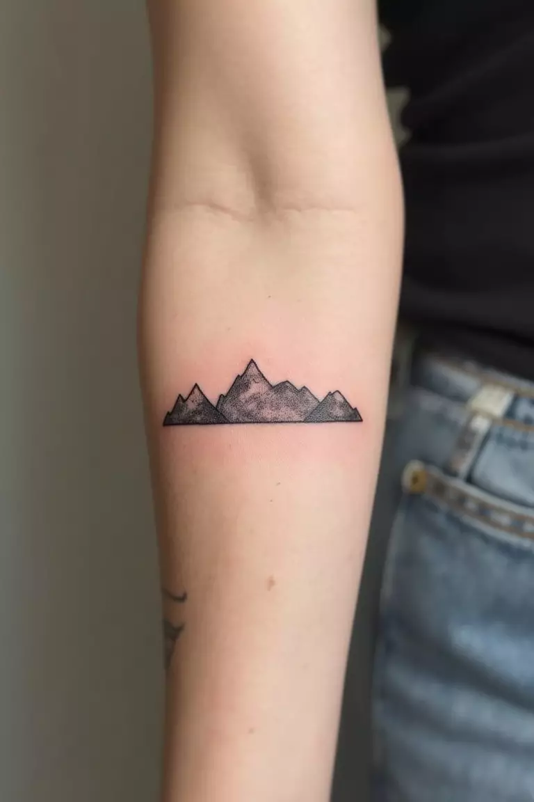 Mountain Range A close-up shot of a woman's arm with a black ink tattoo of a minimalist mountain range outline.